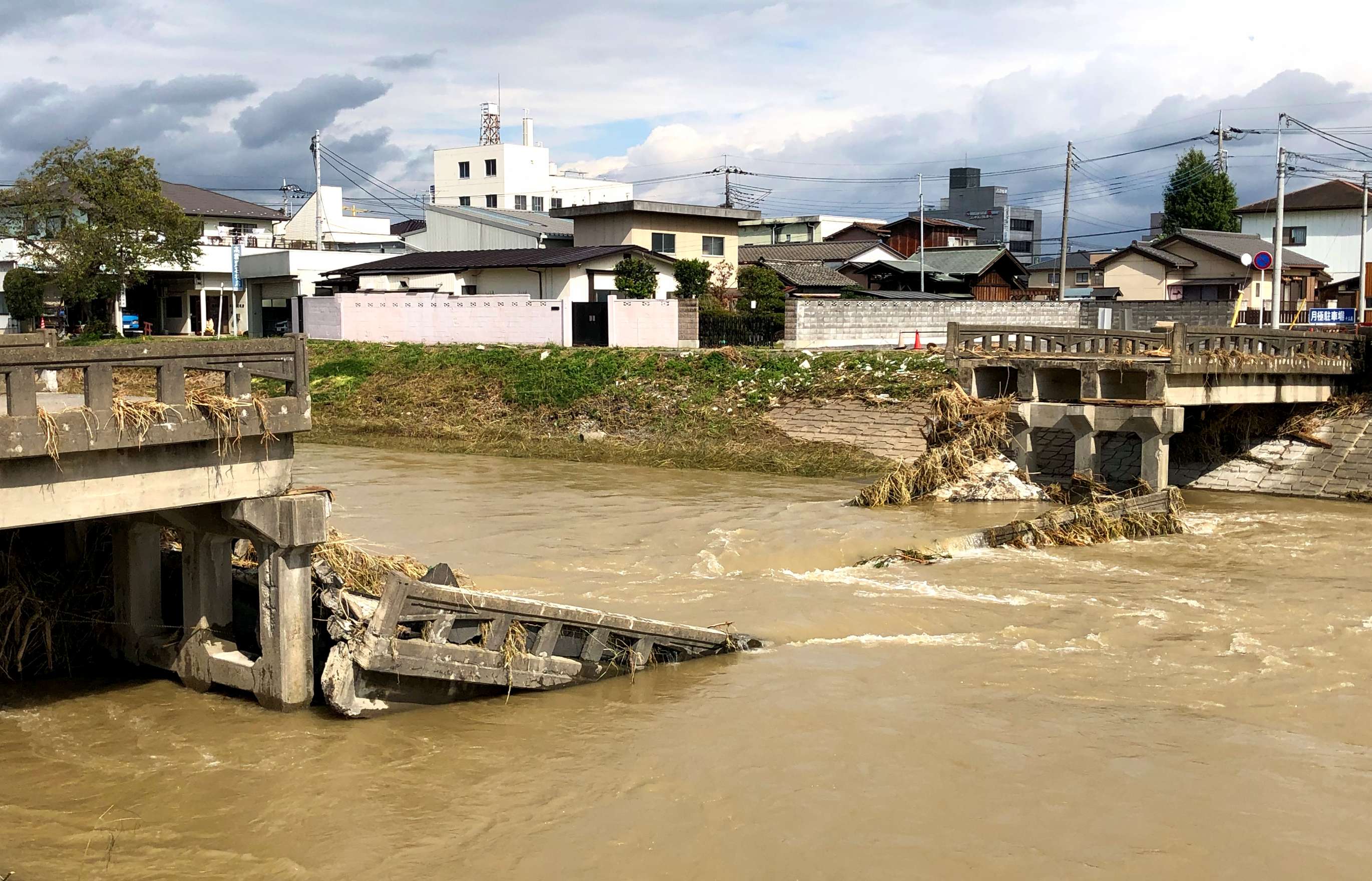 A photograph of a bridge collapsed into a river of fast moving, muddy water with houses and rooftops of a town visible in the background.