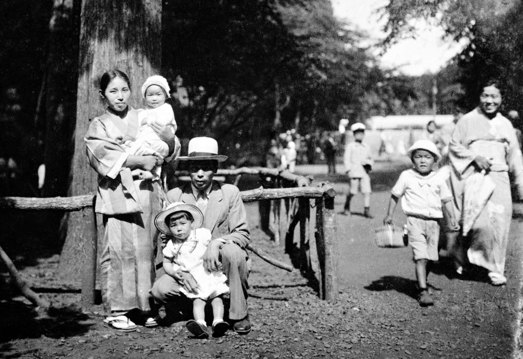Tomoji in kimono stands in front of a tree in the park holding an infant as Shinjo in a wide brimmed hat kneels next to her with a toddler; smiling families walk nearby.