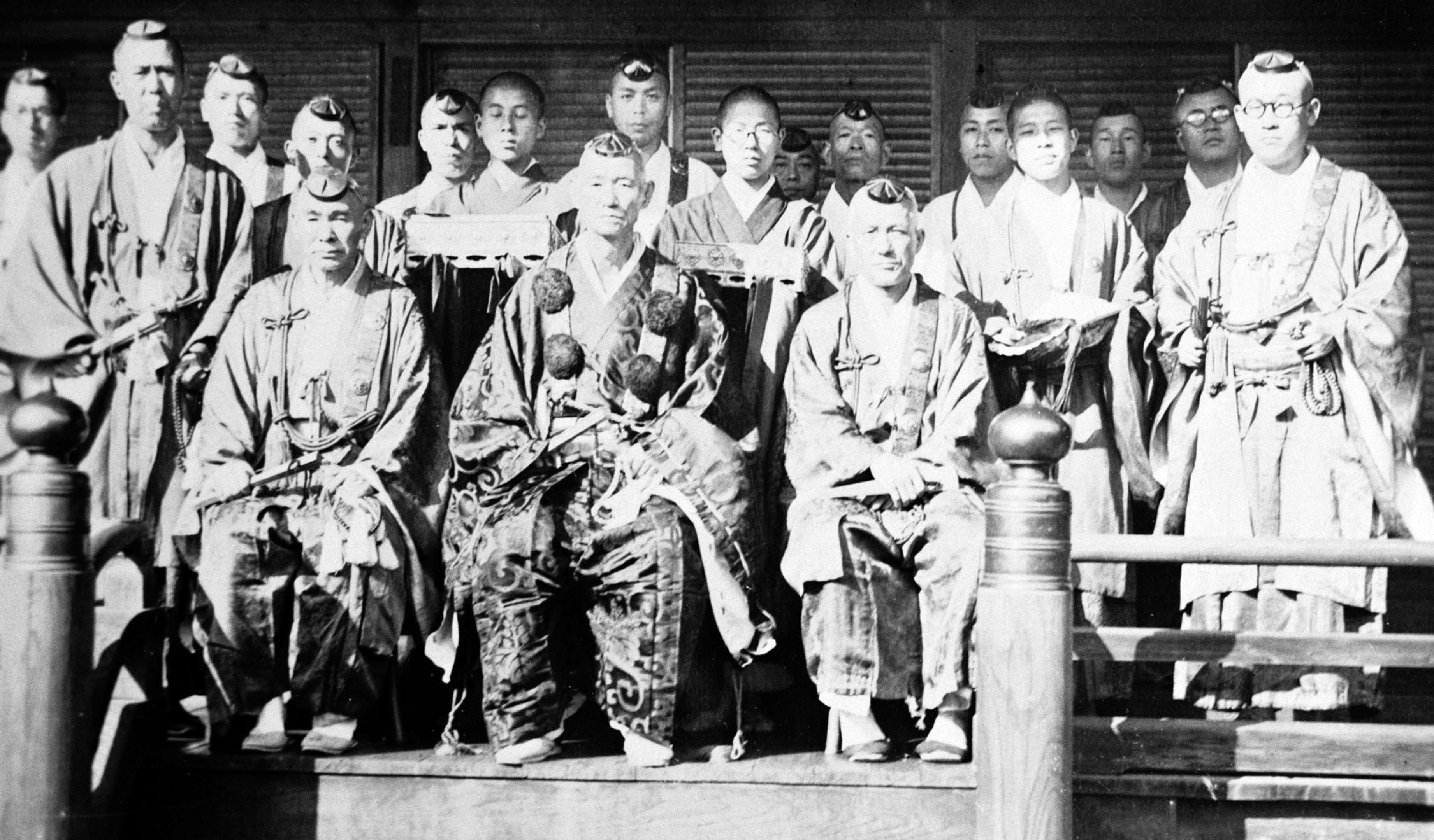 A group of Buddhist priests, 3 seated, 14 standing, all wearing robes, pose for a portrait on a temple portico; the central figure wears elaborate brocade robes adorned with pom-poms.