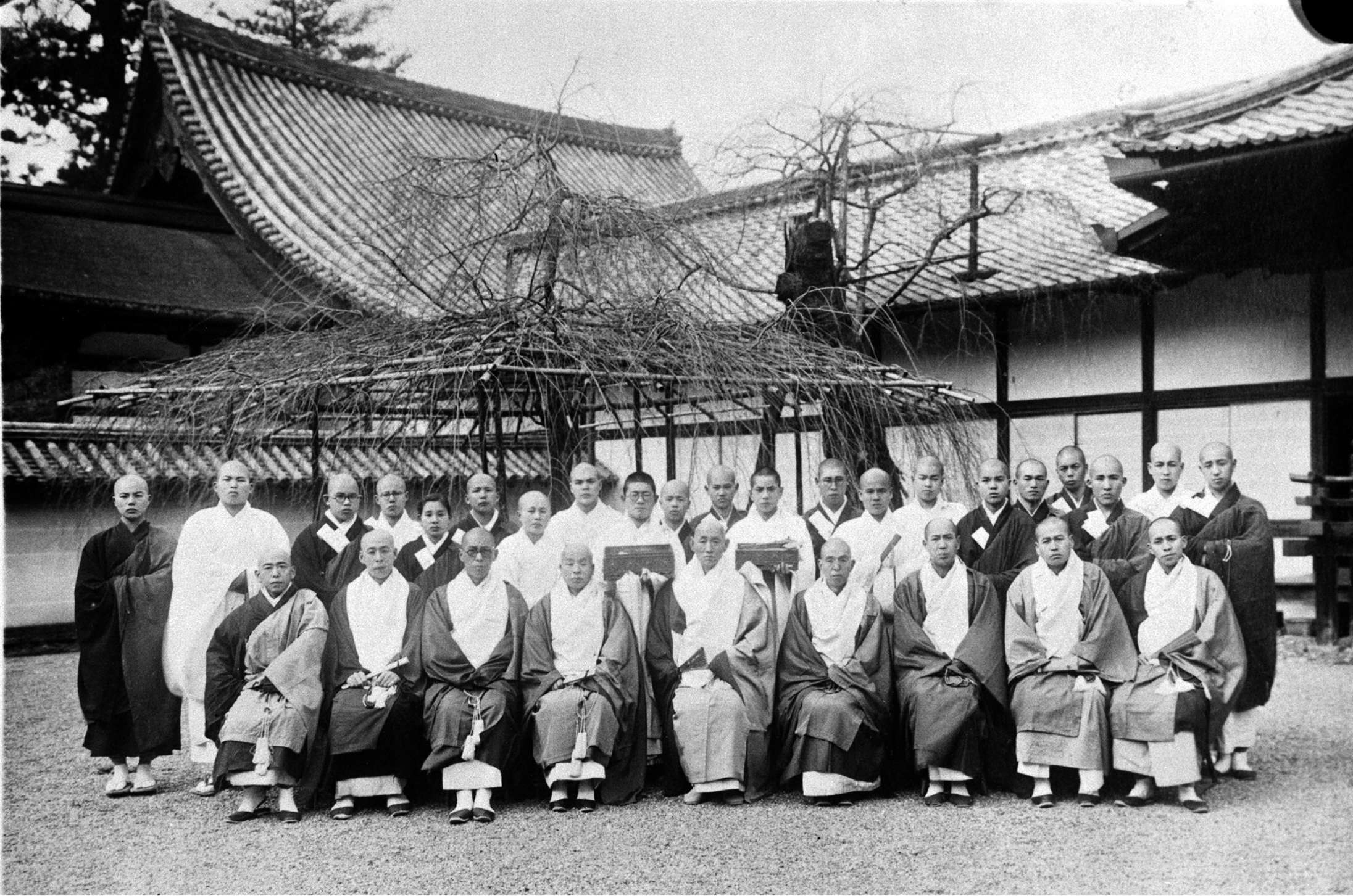 A group of Buddhist priests wearing formal robes, some seated, the rest standing behind, pose for a portrait in the graveled courtyard of a large temple with tiled roofs.