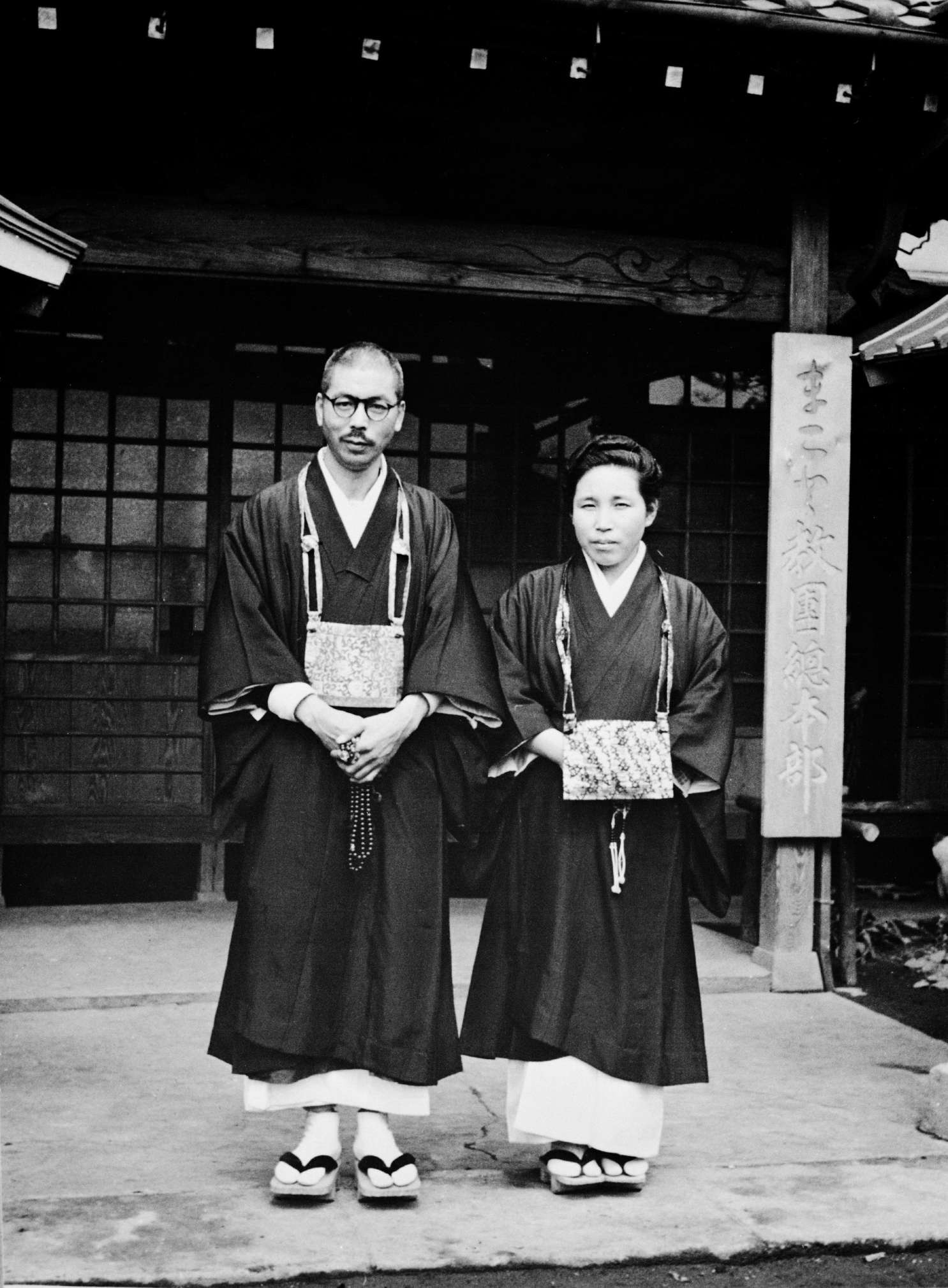 Shinjo and Tomoji wearing priestly robes stand at the entrance to Shinchoji temple; a pillar bearing Japanese calligraphy is visible to the right.