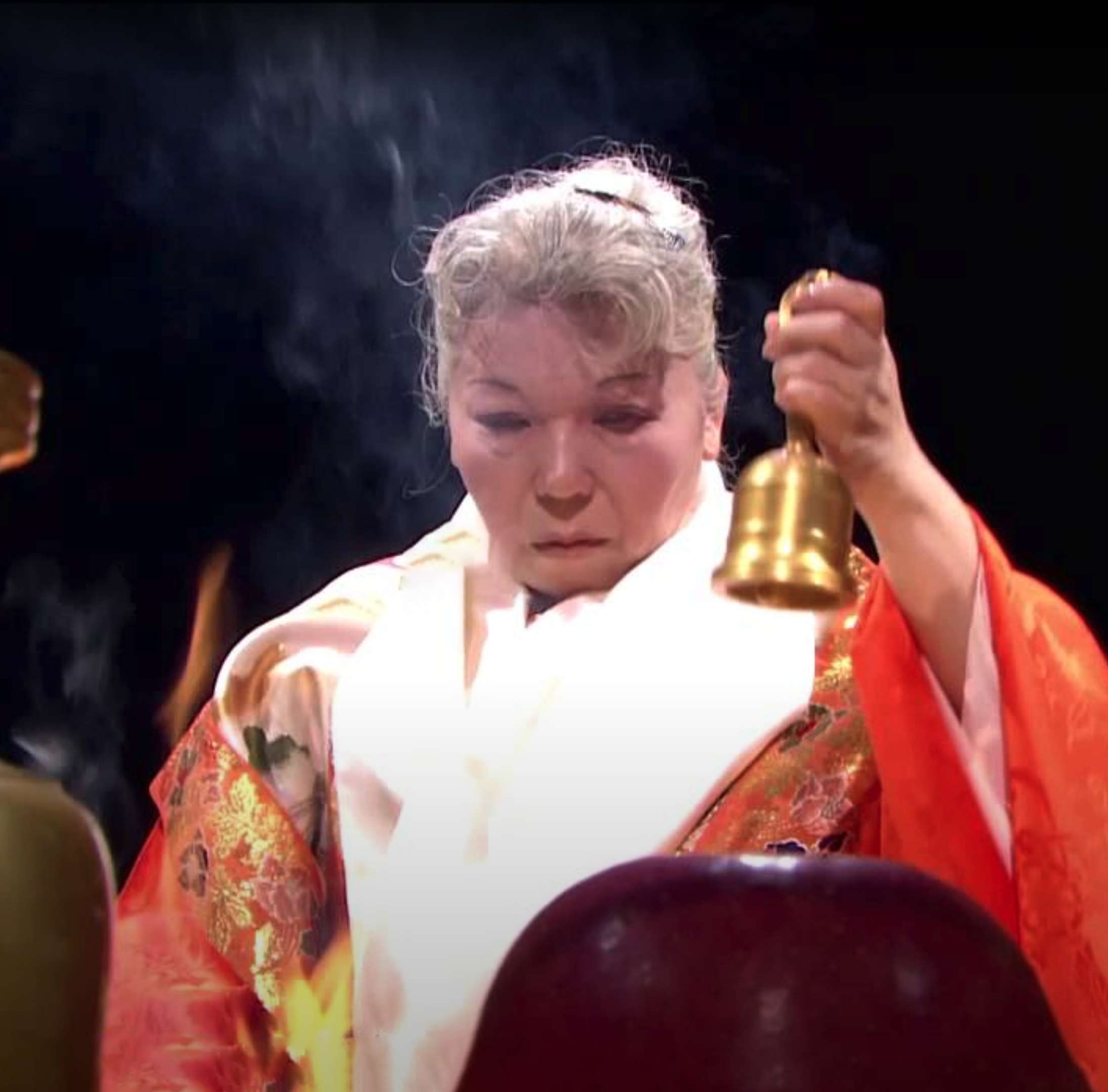 Her Holiness, in bright orange and white robes, rings a ritual hand bell in front of a open fire with an expression of deep concentration.