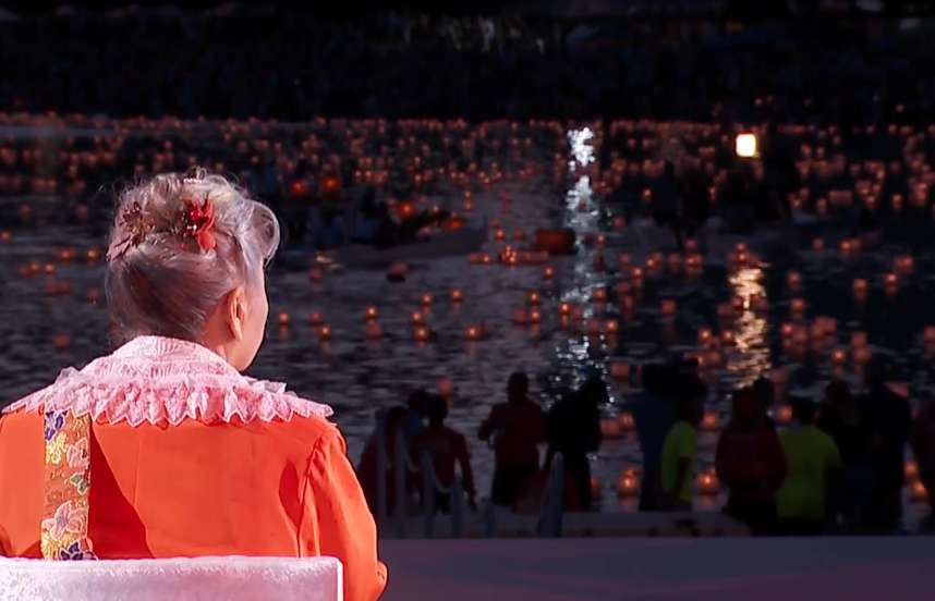 Her Holiness, wearing a red gown, sits watching individuals float lanterns as the sun goes down on the ocean.