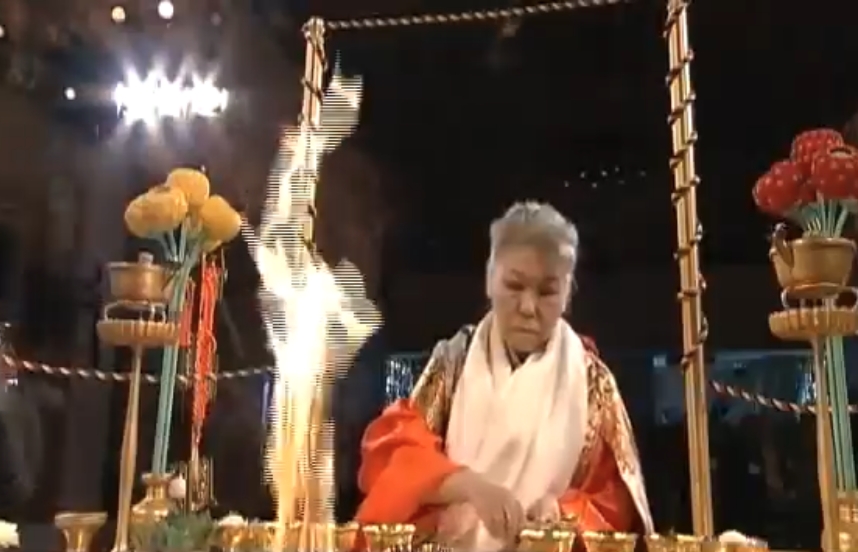 A still image from a video of Her Holiness in ceremonial robes performing a fire ceremony at an altar; a large flame can be seen in the foreground.